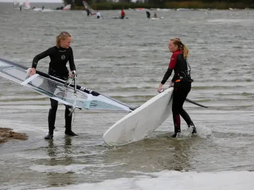 Zwei Mädchen bereiten sich auf das Windsurfen am IJsselmeer vor, tragen Neoprenanzüge und halten ihre Ausrüstung.