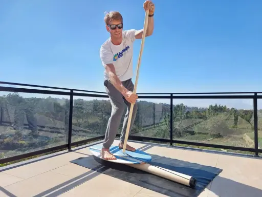 Mann übt Balance-Board-Training zuhause auf einer Terrasse. Er hält einen Paddelstock und balanciert auf einem Brett.