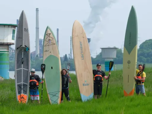 Fünf Personen mit SUP-Boards und Paddeln vor einer Industrieanlage. Die Boards zeigen fortgeschrittene SUP-Technik.