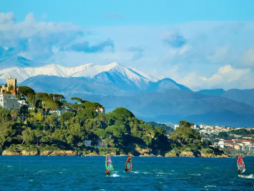 Windsurfen an der französischen Küste mit schneebedeckten Bergen im Hintergrund. Ein Schloss thront auf einem Hügel.