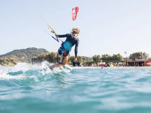 Eine Frau surft mit einem Kite auf Kos. Sie fährt auf einer Welle, Gischt spritzt. Im Hintergrund ist der Strand mit Bäumen und einem Gebäude zu sehen.