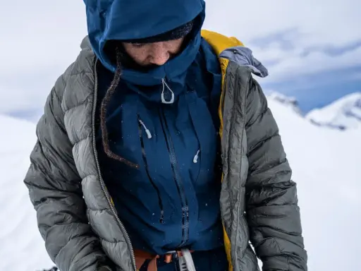 Person in Daunenjacke im Schnee. Die Jacke ist grau und blau, mit gelben Akzenten. Perfekt für das Daunenjacke waschen nach einer Bergtour.