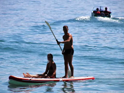Ein Mann steht auf einem Stand Up Paddle Board auf dem Meer, während eine Frau sitzt. Im Hintergrund fährt ein Boot.