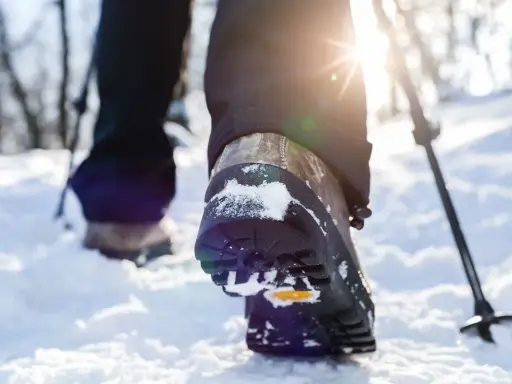 Schneeschuhwandern Schuhe mit Schnee bedeckt, während die Sonne durch die Bäume scheint. Ein Wanderstock ist im Hintergrund zu sehen.