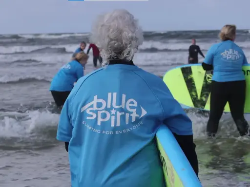Ältere Dame mit grauem Haar und Surfbrett, bereit für Hvide Sande Surfen. Mehrere Personen in blauen Shirts mit "Blue Spirit" Aufdruck im Wasser.