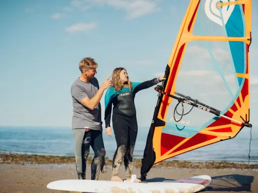 Vater erklärt Tochter das Windsurfen NRW. Sie stehen am Strand mit einem Segel und Board.