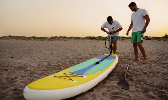 Zwei Männer bereiten ein SUP-Board am Strand vor. Einer pumpt es auf, fragt sich: "Sup, wieviel Bar?
