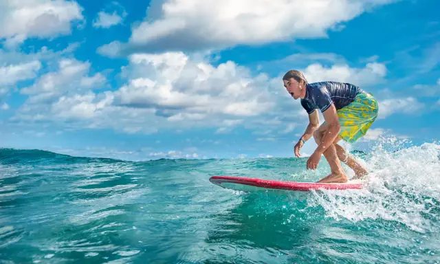 Ein Surfer reitet eine Welle vor der Küste der Dominikanischen Republik. Der Mann trägt ein blaues Shirt und bunte Badeshorts.