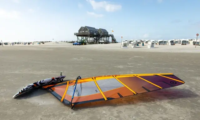 Windsurfen St. Peter-Ording: Segel und Brett liegen am Strand, bereit für den nächsten Ritt auf den Wellen. Strandkörbe und das markante Stelzenhaus im Hintergrund.