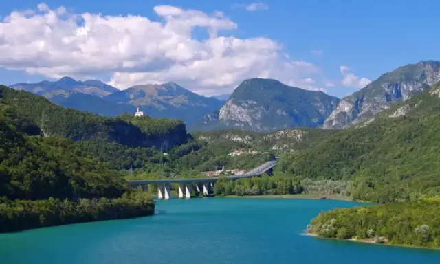 Türkises Wasser des Lago di Cavazzo umgeben von grünen Bergen. Eine Brücke überspannt den See, im Hintergrund eine kleine Kirche auf einem Hügel.