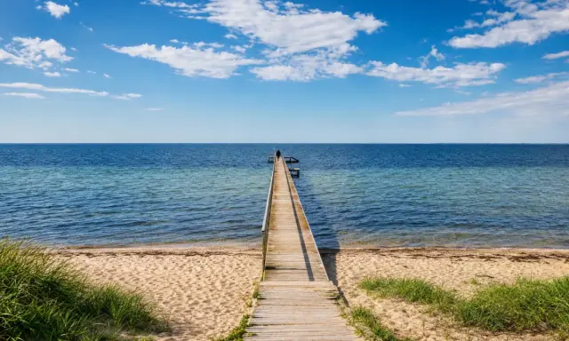 Holzsteg führt ins Meer am Ringkøbing Fjord. Eine Person steht am Ende des Stegs. Blauer Himmel mit Wolken.