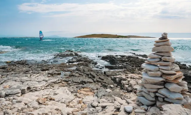 Ein Windsurfer gleitet über das türkisfarbene Meer vor Kroatien. Steinformationen säumen die felsige Küste.
