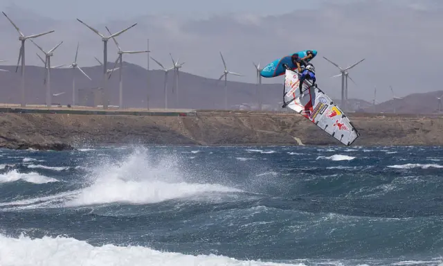Windsurfer Britt Dunkerbeck springt hoch über den Wellen, im Hintergrund Windräder.
