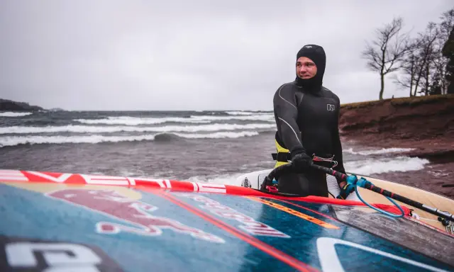 Mann in Neoprenanzug mit Windsurfbrett am Strand. Kaltes Wetter für Windsurfen in Deutschland.