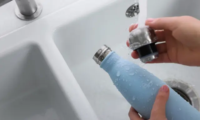 Hände reinigen eine hellblaue Trinkflasche unter fließendem Wasser im Waschbecken.
