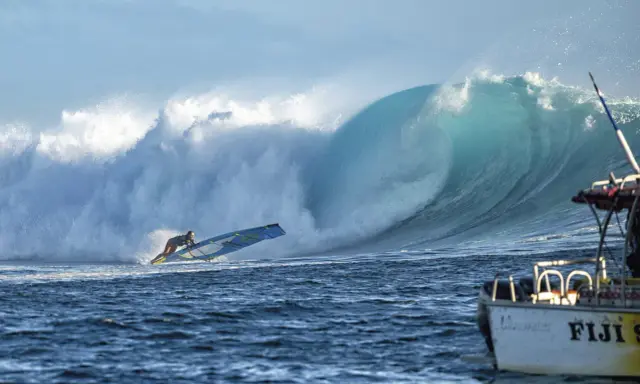 Sarah Hauser surft auf einer riesigen Welle, die sich hinter ihr bricht. Ein Boot mit der Aufschrift "Fiji S" liegt im Vordergrund.
