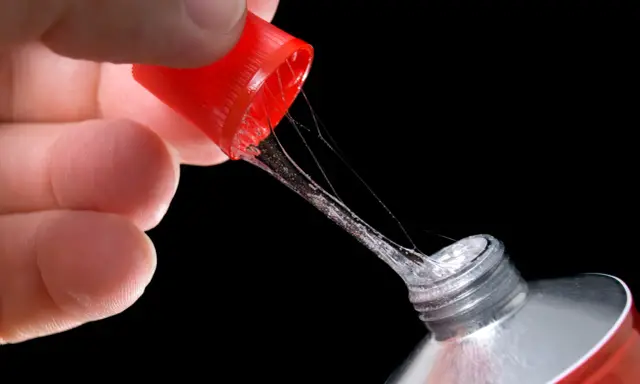 Ein Finger hält die rote Kappe eines Tubenklebers, aus dem zäher, glitzernder Kleber in Fäden heraustropft.