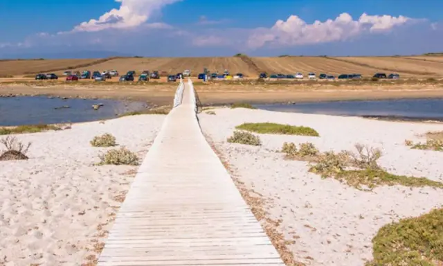 Holzsteg führt über Sand zu einem ruhigen Gewässer. Dahinter parken Autos, die auf einen Ausflug am **mari ermi** warten.