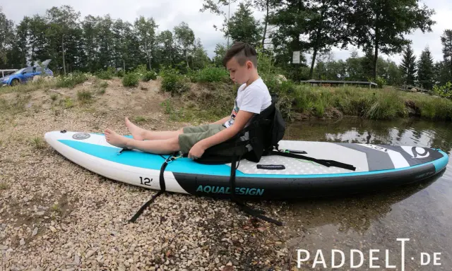 Junge sitzt auf SUP-Board, um den SUP Sitz zu befestigen. Das Board liegt am Ufer.