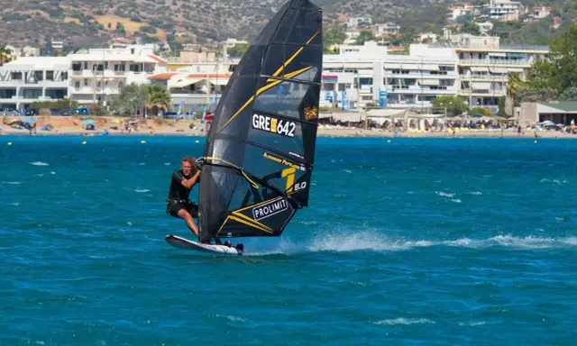 Windsurfer mit schwarzem Segel und gelben Akzenten auf türkisfarbenem Wasser vor einer Küste mit Gebäuden und Bergen. Surfen in Griechenland.