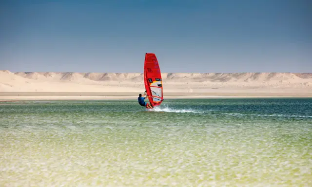 Windsurfer mit rotem Segel gleitet über das Wasser bei Dakhla, Marokko, vor einer Wüstenlandschaft.