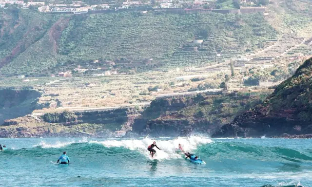 Surfer auf La Gomera reiten eine Welle, während im Hintergrund terrassierte Hänge und Häuser zu sehen sind.