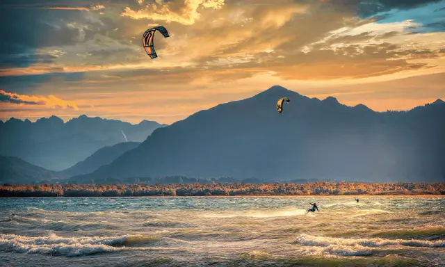 Kitesurfer gleiten über die Wellen am Chiemsee, während die Sonne hinter den Bergen untergeht. Ein traumhafter Abend für Windsurfen.