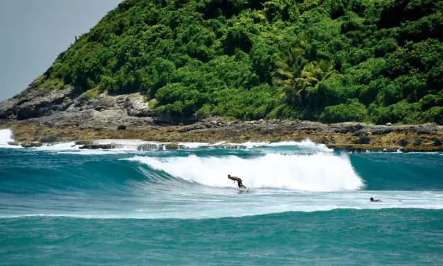 Ein Surfer reitet eine Welle vor der üppig grünen Küste von Guadeloupe. Das türkisfarbene Wasser und die felsige Küstenlinie bieten die perfekte Kulisse für das Surfen.