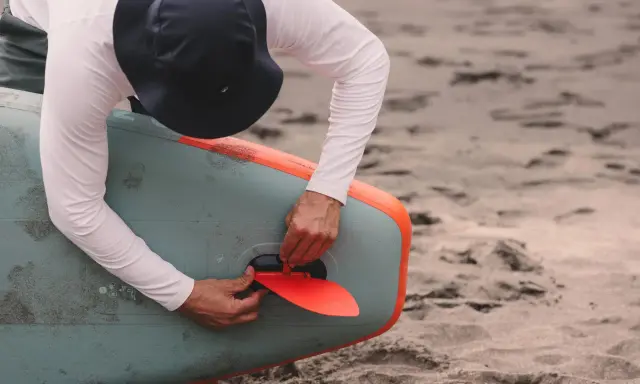 Mann mit Hut befestigt die rote Finne an einem aufblasbaren SUP-Board. Gute SUP-Pflege beginnt mit dem richtigen Zubehör.