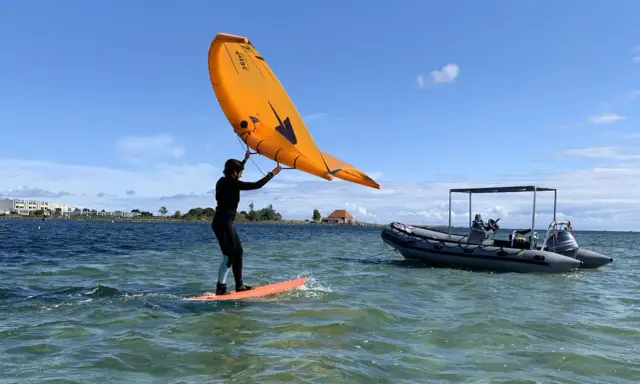 Person auf einem Board mit einem orangen Flügel, der über dem Wasser schwebt. Ein Schlauchboot liegt daneben. Foil surfen ohne Segel.