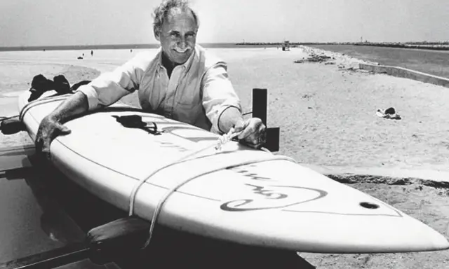 Jim Drake, ein Pionier des Surfens, lächelt stolz neben seinem Surfbrett am Strand.