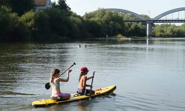 Zwei Mädchen genießen das Stand Up Paddling in Heidelberg. Im Hintergrund eine Brücke und Bäume.