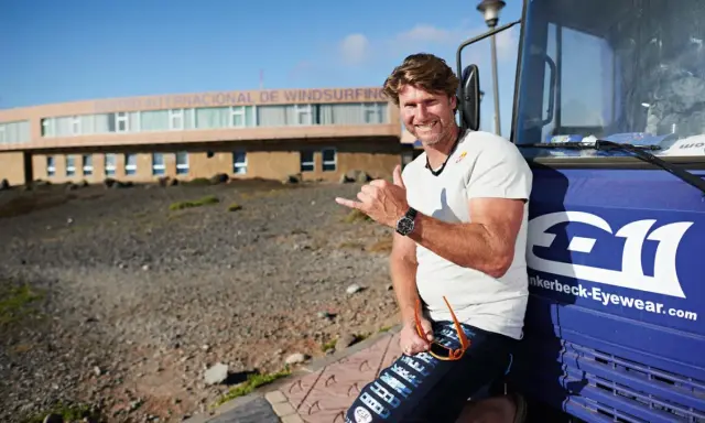 Bekannte deutsche Windsurfer: Ein Mann mit Sonnenbrille und Shaka-Geste lehnt an einem blauen Bus vor dem Centro Internacional de Windsurfing.