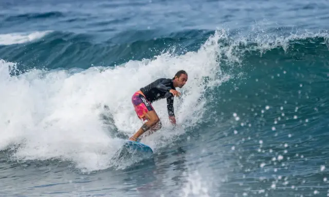 Ein Surfer reitet eine Welle vor Zypern. Er trägt ein schwarzes Rashguard und bunte Boardshorts.