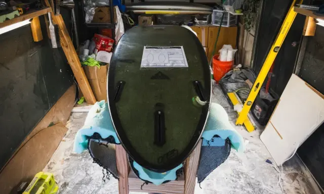 Surfboard liegt auf Ständer, bereit für die **Surfboard Reparatur**. Werkzeuge und Materialien im Hintergrund.
