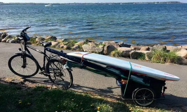 Fahrradanhänger mit Surfbrett und Paddel am Seeufer, bereit für ein Abenteuer auf dem Wasser.