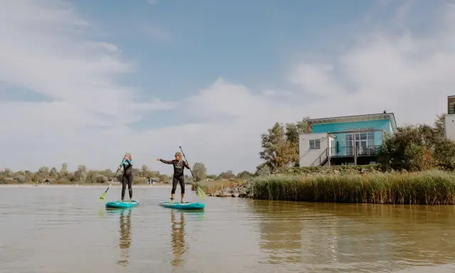 Zwei Frauen auf SUP-Boards am Makkum Strand, mit einem modernen Haus im Hintergrund.