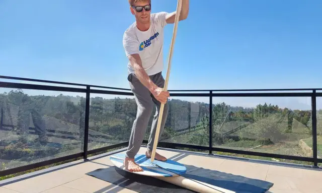 Mann übt Balance-Board-Training zuhause auf einer Terrasse. Er hält einen Paddelstock und balanciert auf einem Brett.
