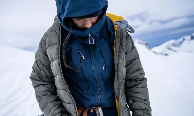 Person in Daunenjacke im Schnee. Die Jacke ist grau und blau, mit gelben Akzenten. Perfekt für das Daunenjacke waschen nach einer Bergtour.