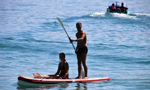 Ein Mann steht auf einem Stand Up Paddle Board auf dem Meer, während eine Frau sitzt. Im Hintergrund fährt ein Boot.