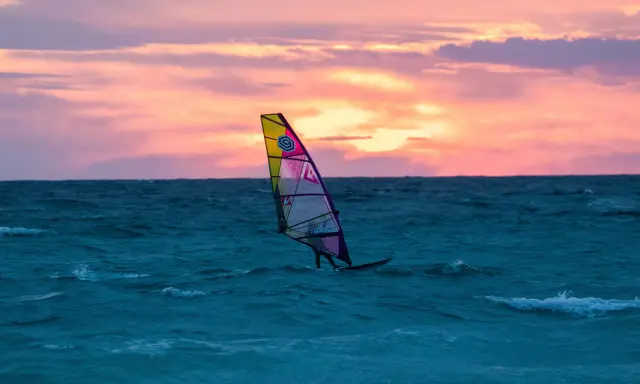 Ein Windsurfer gleitet bei Sonnenuntergang über das Meer. Die bunten Wolken spiegeln sich im Wasser. Ein perfekter Tag für Windsurfen Mallorca.