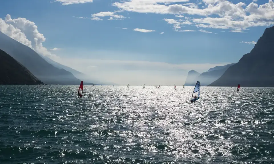 Windsurfer tummeln sich auf dem glitzernden Gardasee vor einer Bergkulisse.