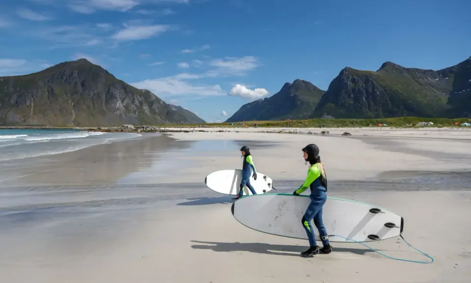 Zwei Surferinnen mit Neoprenanzügen und Surfbrettern am Strand in Norwegen. Die Berge im Hintergrund sind grün und die Wellen sind sanft.