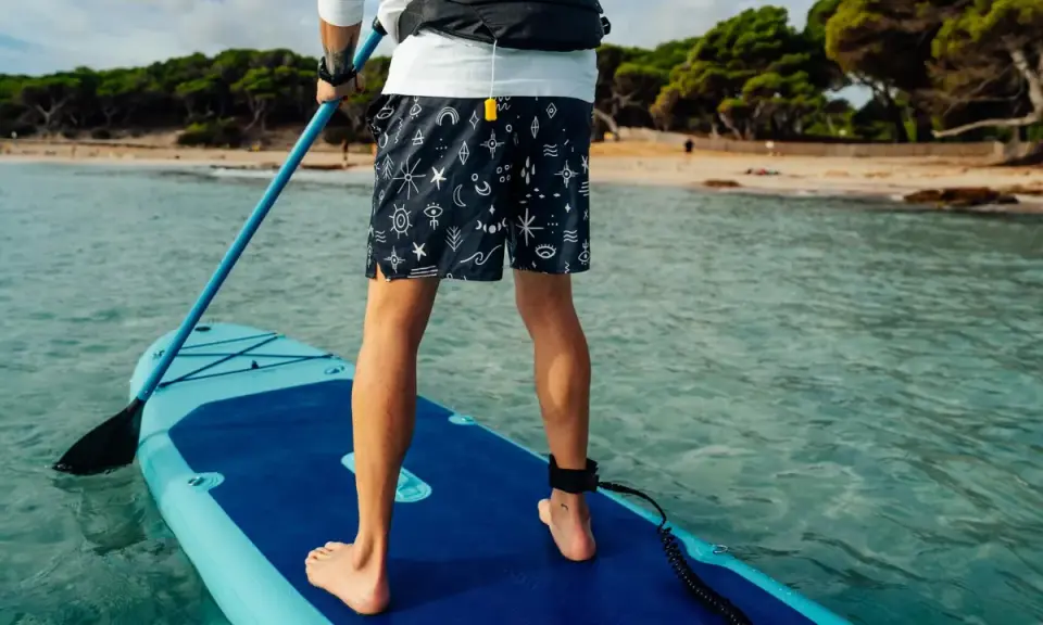 Person übt SUP Training auf einem Board im türkisfarbenen Wasser. Im Hintergrund eine sandige Bucht mit Bäumen.