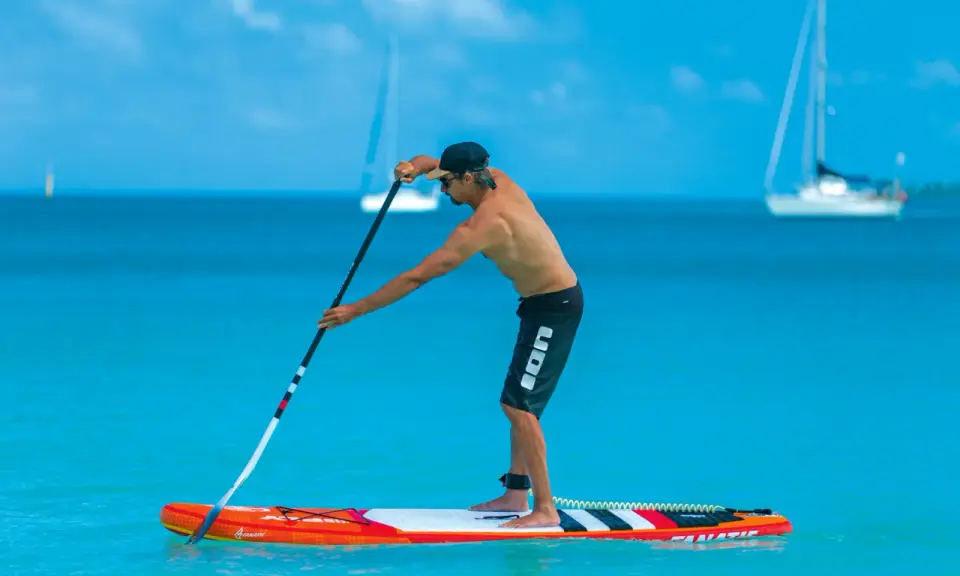Mann stellt sein SUP Paddel ein, während er auf einem Board im türkisfarbenen Wasser steht. Segelboote sind im Hintergrund zu sehen.