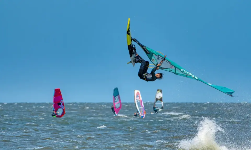 Windsurfer vollführt spektakulären Sprung über den Wellen. Mehrere andere Windsurfer sind auf dem Wasser zu sehen, was auf beliebte Windsurfing Spots hindeutet.
