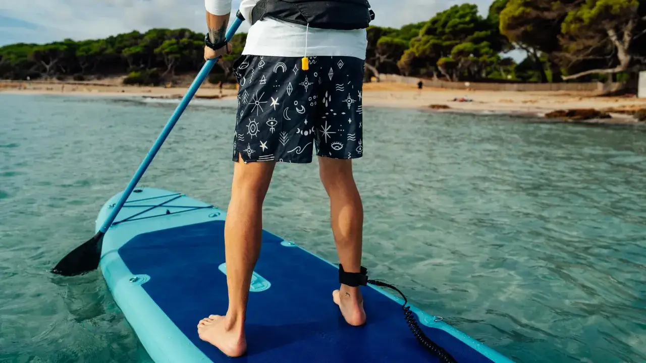Person übt SUP Training auf einem Board im türkisfarbenen Wasser. Im Hintergrund eine sandige Bucht mit Bäumen.