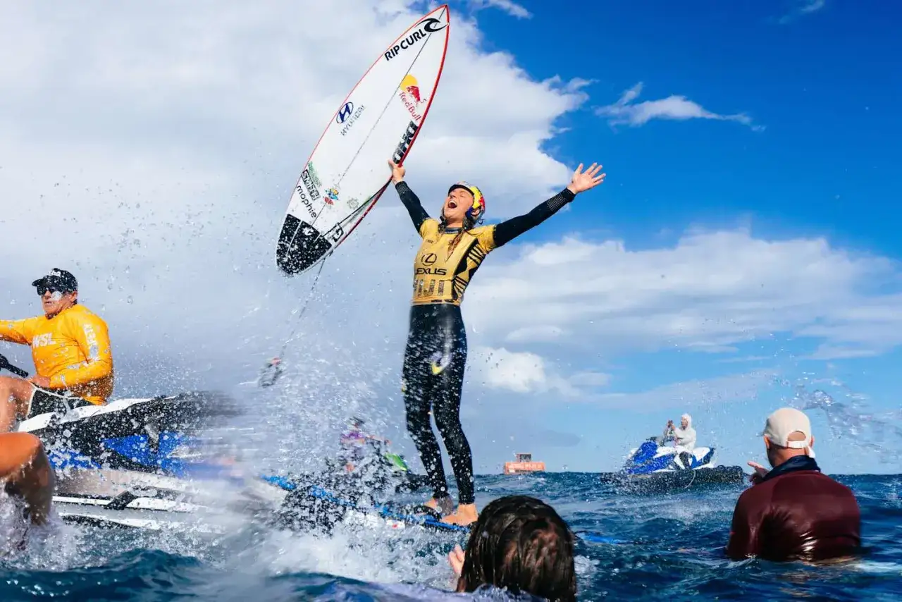 Jubelnder Surfer mit Surfbrett über dem Kopf, umgeben von Wasserspritzern. Er könnte der beste Surfer der Welt sein, gefeiert von den Zuschauern.