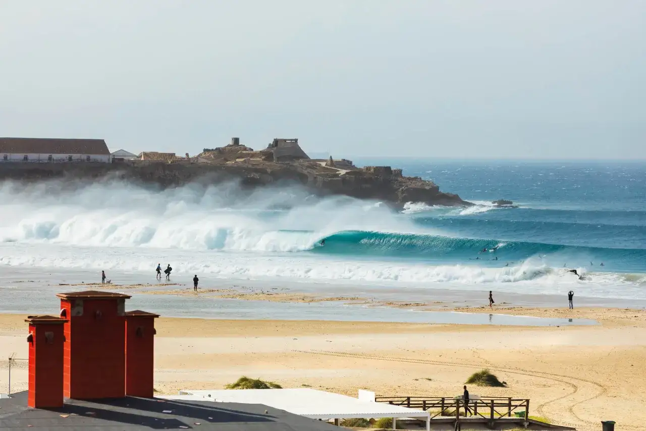 Perfekte Wellen f&uuml;r Tarifa Surfen. Surfer reiten die t&uuml;rkisfarbene Welle vor einer felsigen K&uuml;ste mit Ruinen.