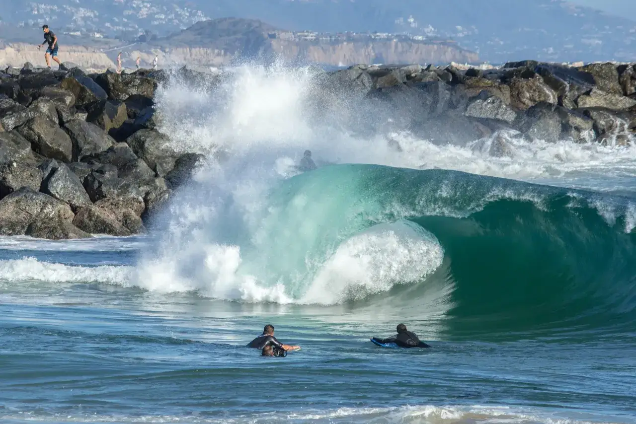 Surfer warten auf den perfekten shore break. Eine riesige, türkisfarbene Welle bricht über Felsen.